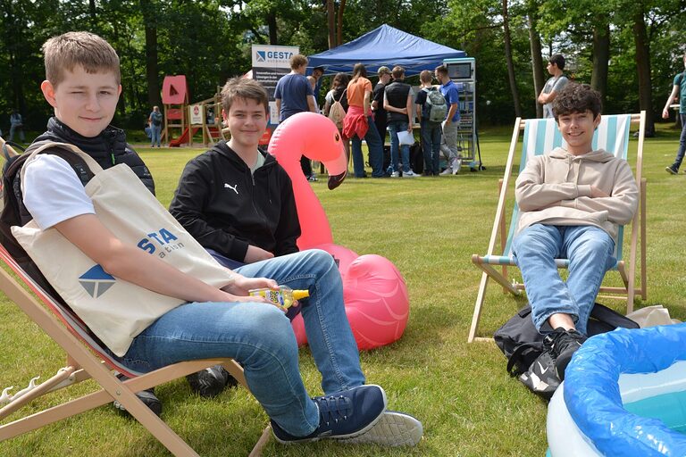 Drei Jungs sitzen in Liegestühlen auf einer Wiese. Im Hintergrund sieht man einen Pavillon, vor dem weitere Schüler stehen.