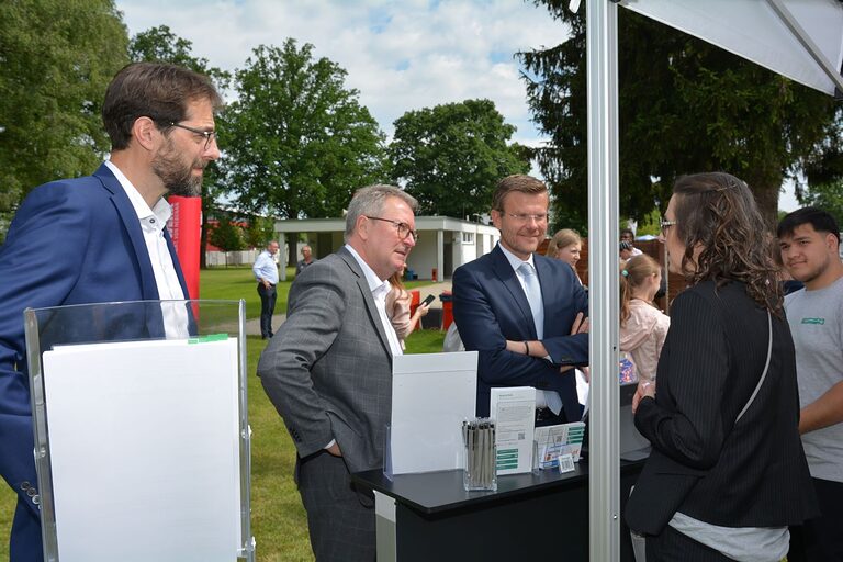 Drei Männer stehen an einem Stand und unterhalten sich mit einer Frau. 