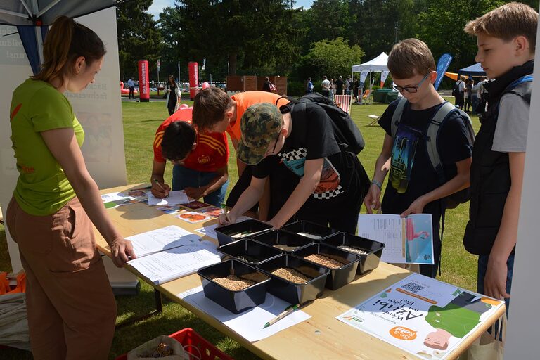 Schüler stehen an einem Stand vor Schalen mit Getreidesorten. Drei füllen ein Quiz aus. Eine Frau beobachtet sie. 