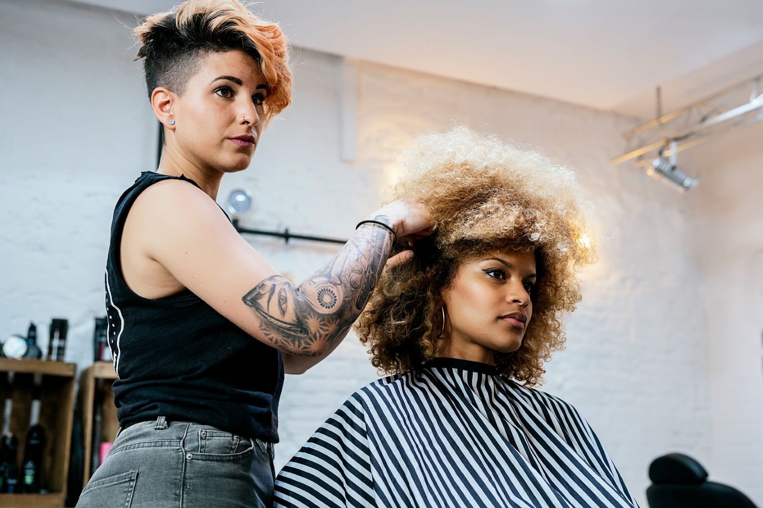 Eine Frau wühlt einer anderen Frau mit sehr vielen Locken in einem Friseursalon in den Haaren. Beide blicken in einen Spiegel