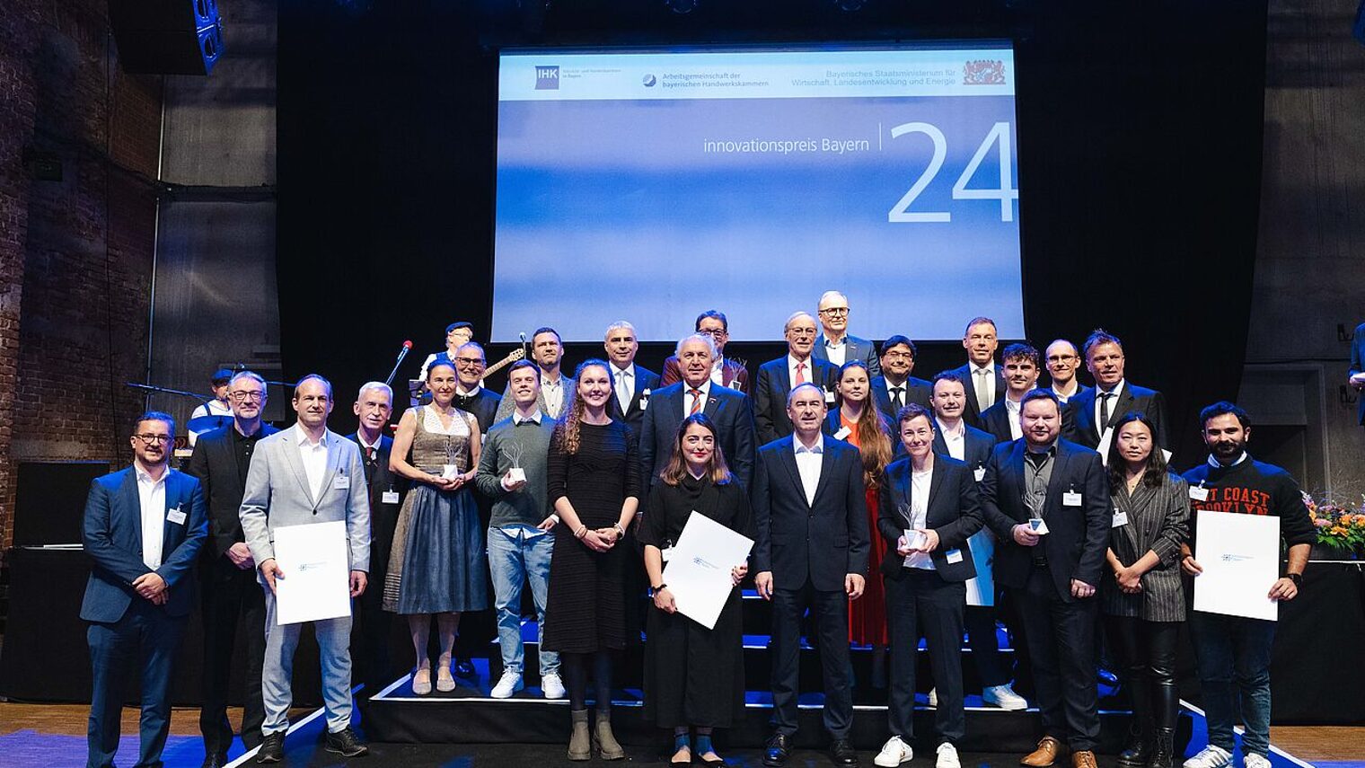 Viele Menschen stehen in festlicher Kleidung vor einer Leinwand, auf der steht: Innovationspreis 24