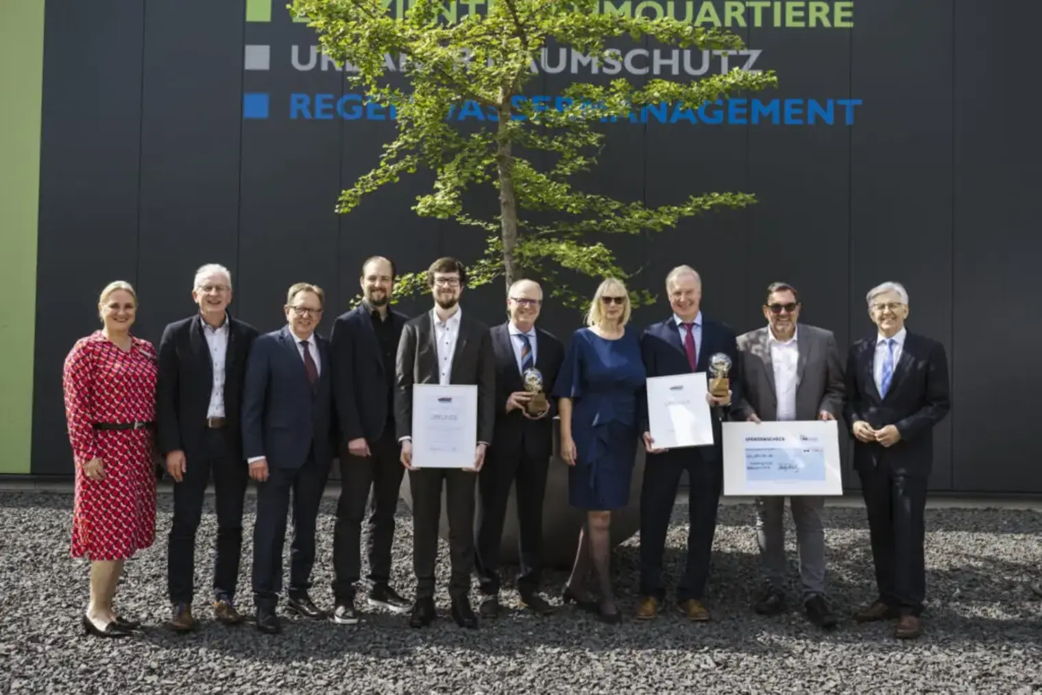 Viele Menschen stehen mit Urkunden vor einem Baum