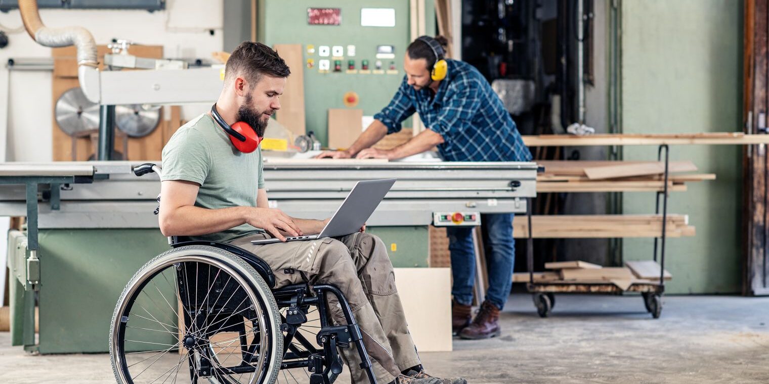 Zwei Handwerker in einer Schreinerwerkstatt: Ein Mann im Rollstuhl arbeitet am Laptop, ein Kollege steht an der Werkbank.