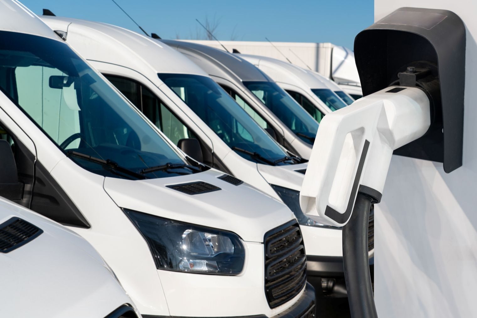 Mehrere weiße Transporter stehen auf einem Betriebsgelände, im Vordergrund eine Wandladestation für Elektrofahrzeuge.