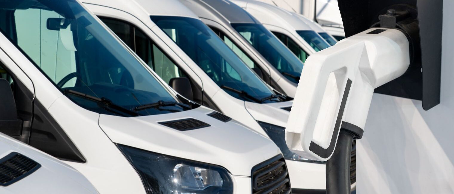 Mehrere weiße Transporter stehen auf einem Betriebsgelände, im Vordergrund eine Wandladestation für Elektrofahrzeuge.