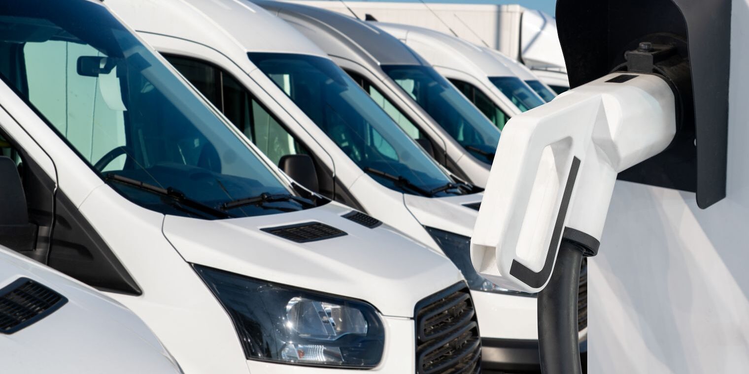 Mehrere weiße Transporter stehen auf einem Betriebsgelände, im Vordergrund eine Wandladestation für Elektrofahrzeuge.