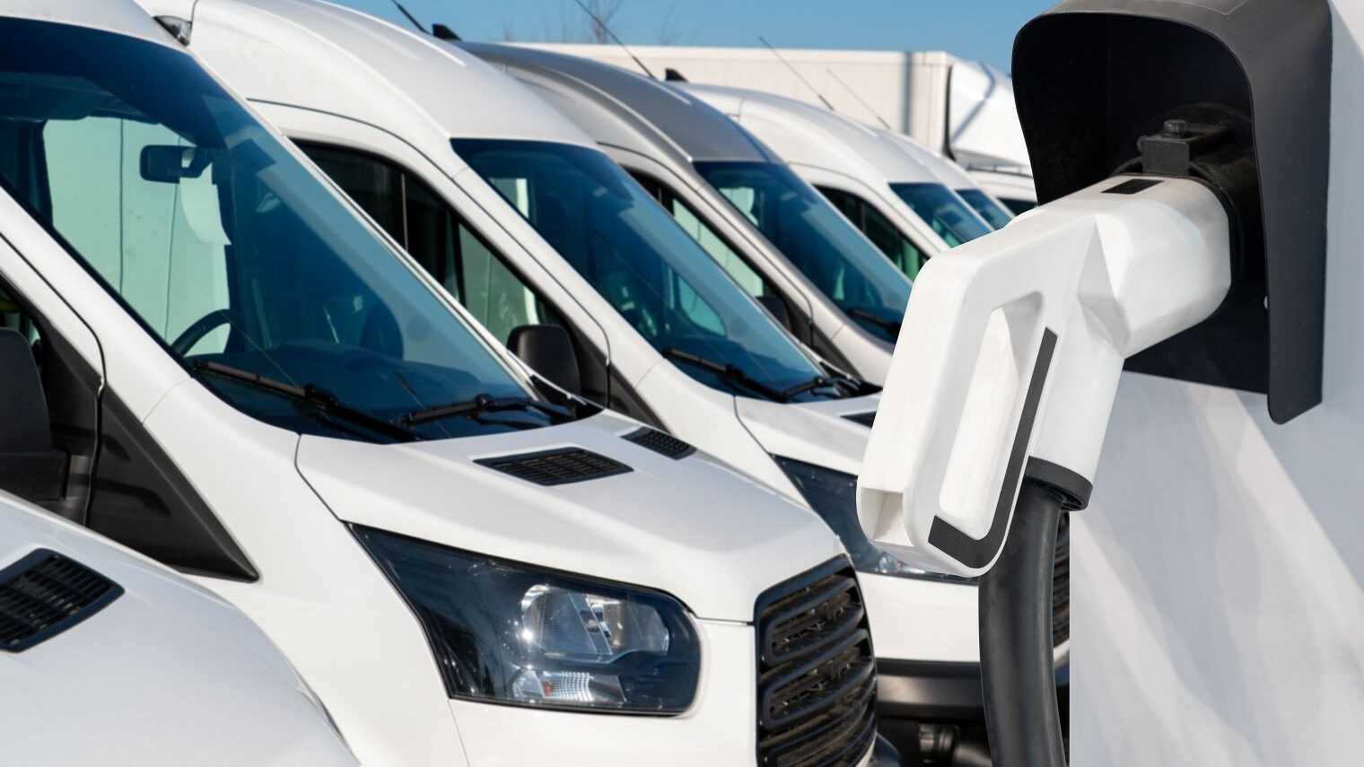 Mehrere weiße Transporter stehen auf einem Betriebsgelände, im Vordergrund eine Wandladestation für Elektrofahrzeuge.