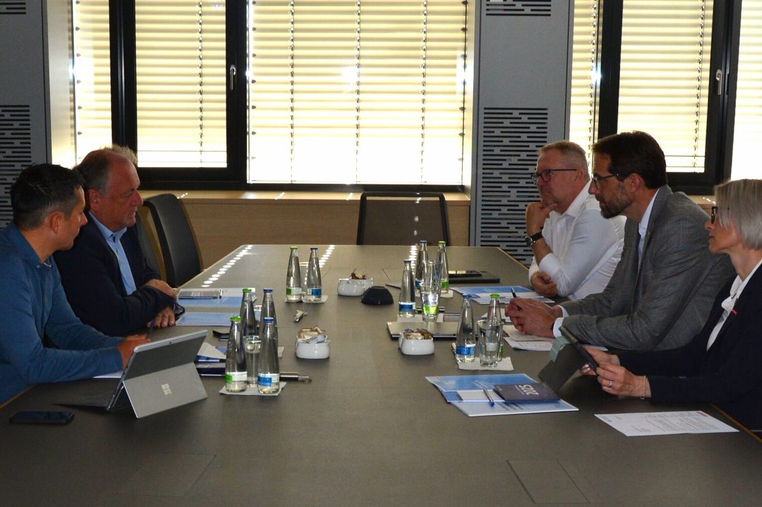 Fünf Personen sitzen an einem Konferenztisch und führen ein Gespräch. Auf dem Tisch liegen Unterlagen und Wasserflaschen.