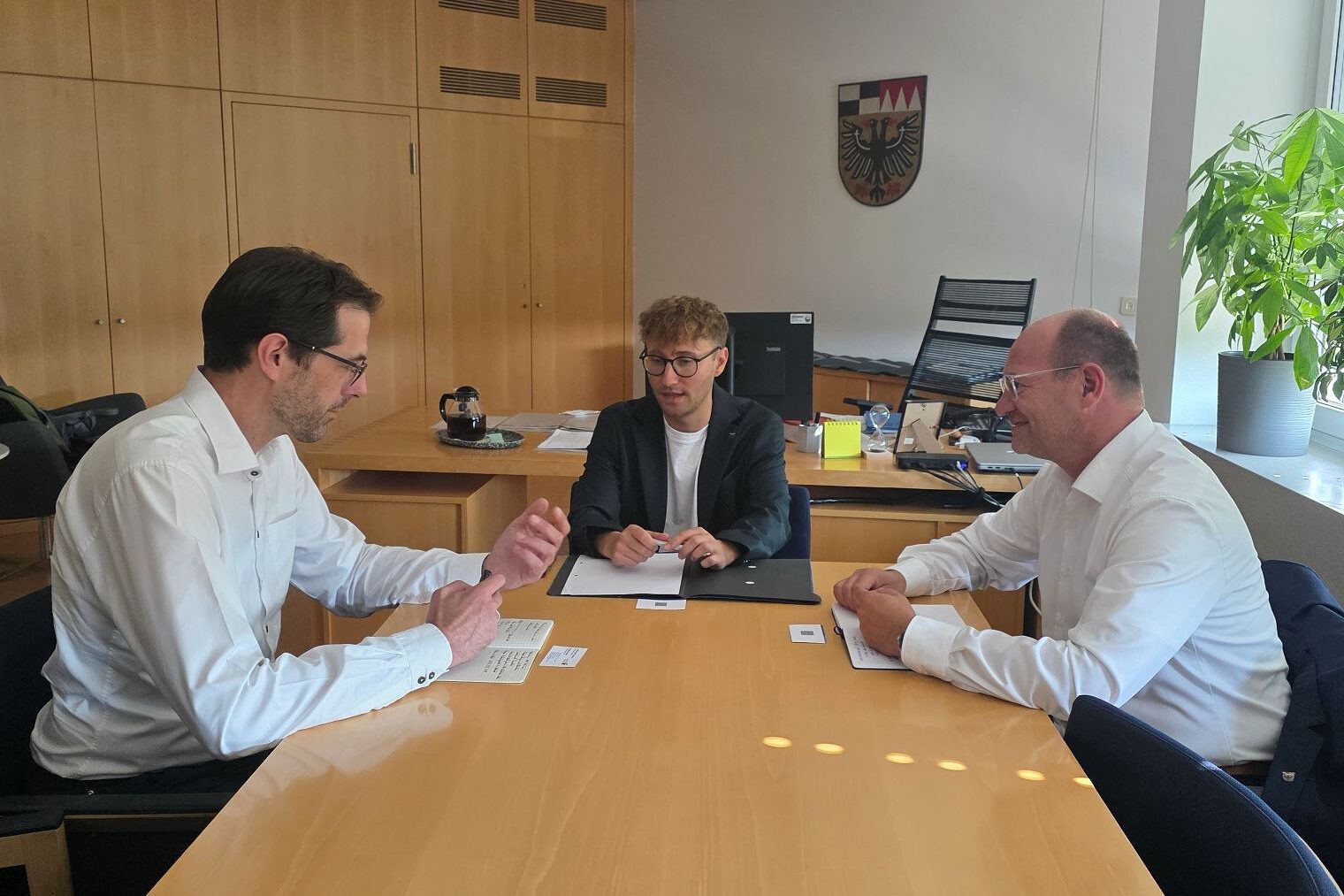 Drei Männer sitzen an einem Besprechungstisch. Im Hintergrund das Wappen des Landkreises Ansbach.