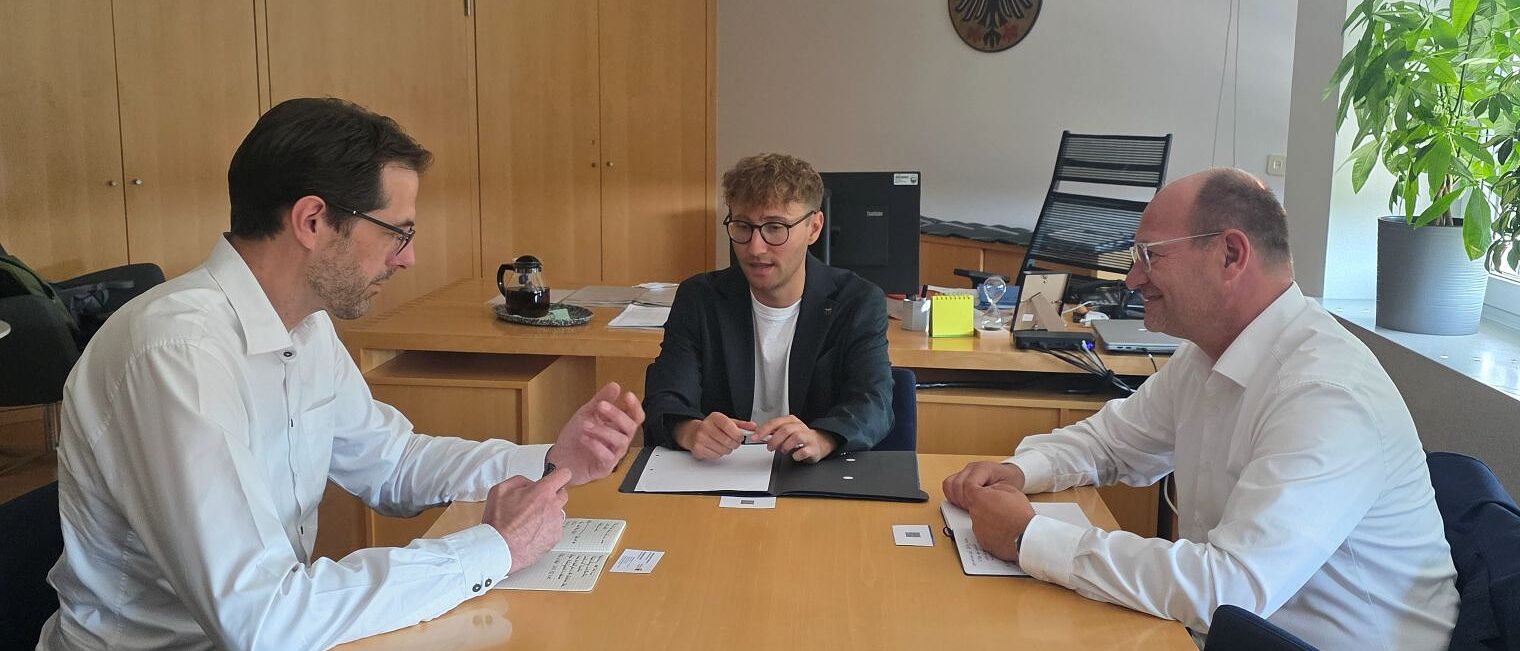 Drei Männer sitzen an einem Besprechungstisch. Im Hintergrund das Wappen des Landkreises Ansbach.