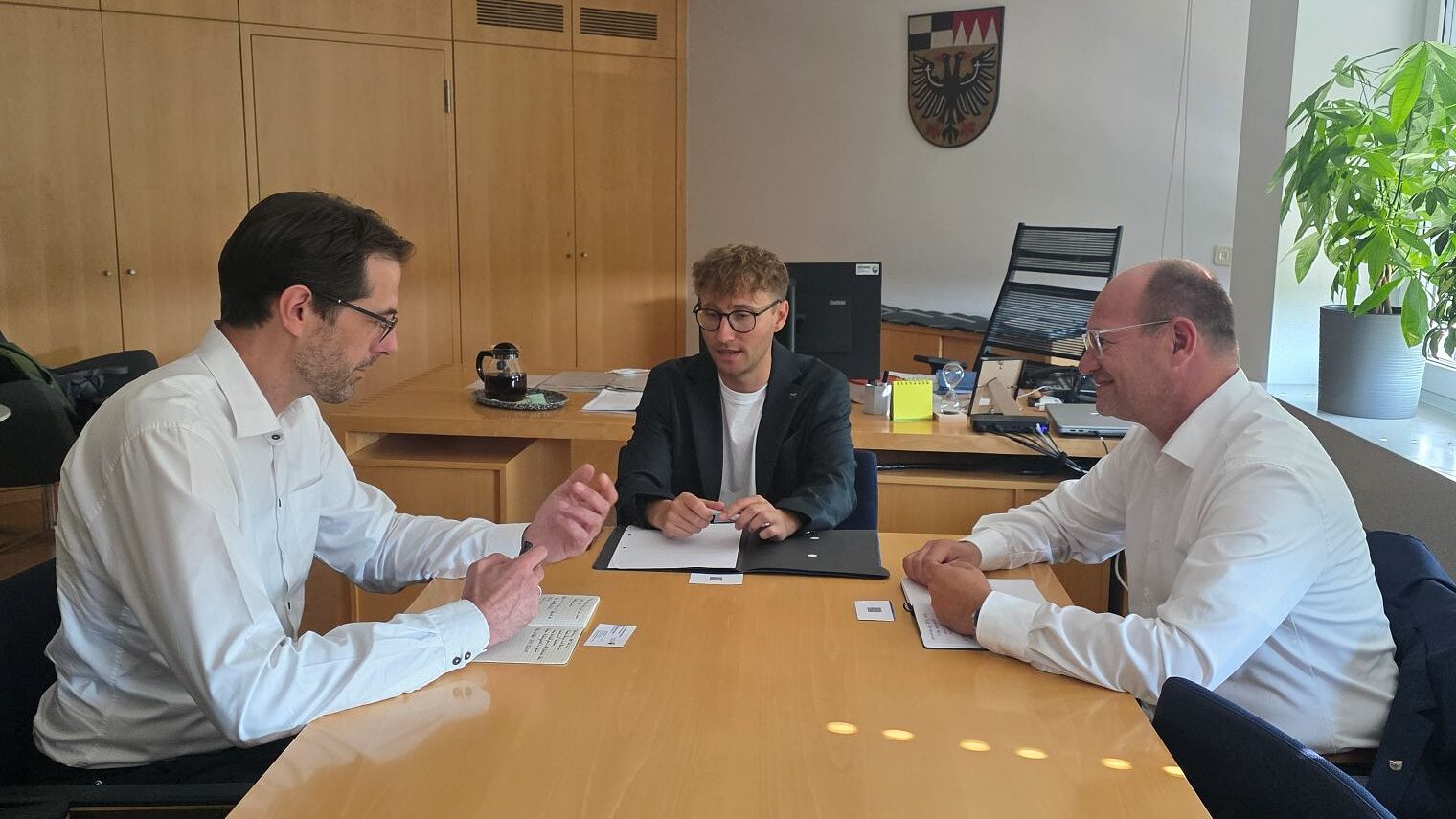 Drei Männer sitzen an einem Besprechungstisch. Im Hintergrund das Wappen des Landkreises Ansbach.