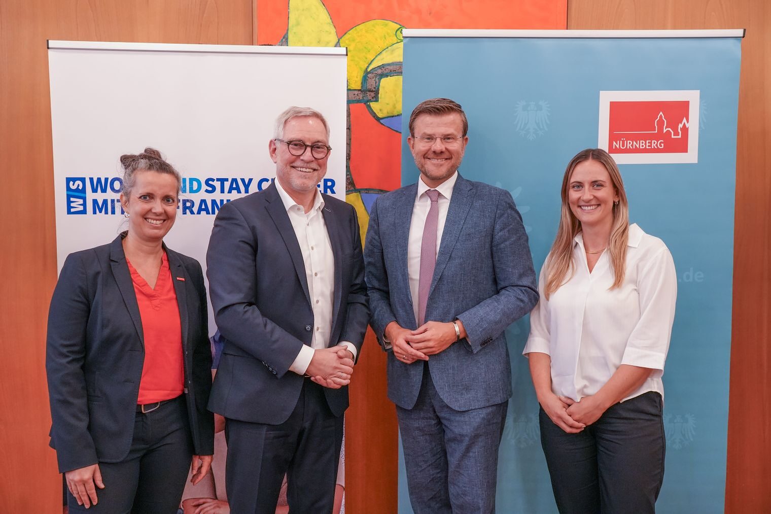 Vier Personen lächeln vor Rollups von Work-and-Stay-Center und Stadt Nürnberg zur Vertragsunterzeichnung.