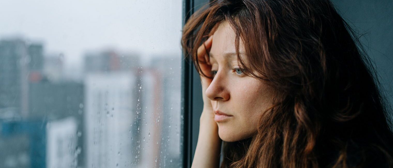 Eine junge Frau blickt nachdenklich durch ein verregnetes Fenster auf die Stadt.
