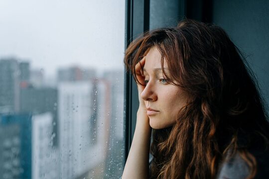 Eine junge Frau blickt nachdenklich durch ein verregnetes Fenster auf die Stadt.