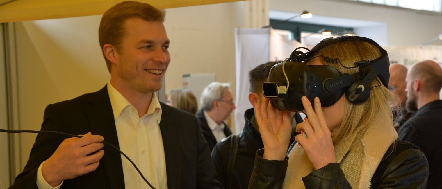 Eine Frau probiert eine VR-Brille aus; ein Mann erklärt ihr das Gerät und hält das Verbindungskabel.