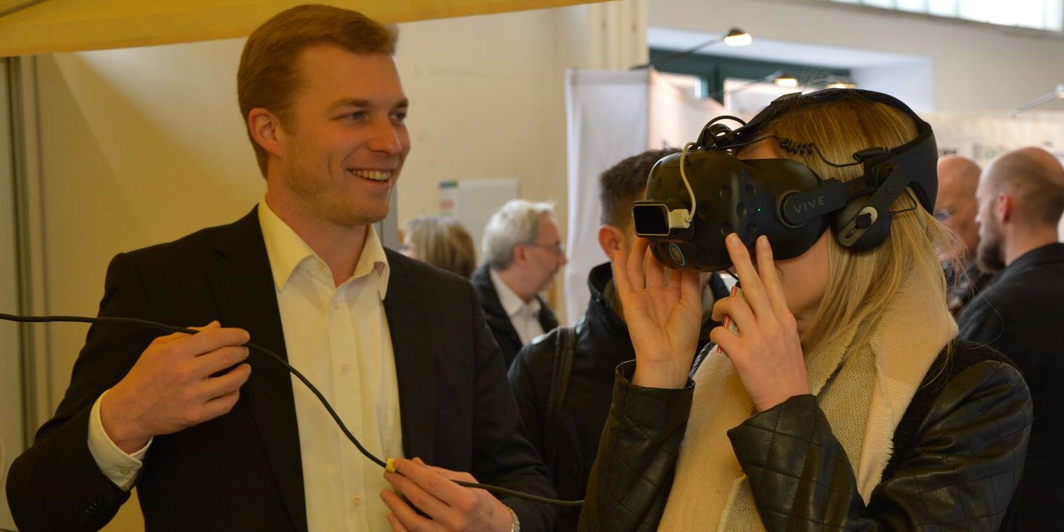 Eine Frau probiert eine VR-Brille aus; ein Mann erklärt ihr das Gerät und hält das Verbindungskabel.