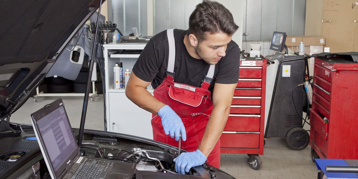 Kfz-Mechatroniker prüft den Motorraum eines Autos und nutzt ein Diagnosegerät in der Werkstatt.